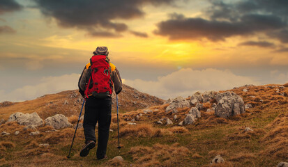 Obraz premium Hiker with a red backpack walking on a rocky mountain trail at sunset, enjoying nature and solitude in the wilderness.