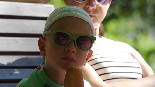 A young boy wearing sunglasses and a headscarf eats a breadstick while sitting next to his mother. They relax on a wooden bench surrounded by green trees on a bright day.
