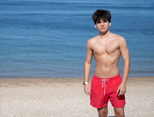 Portrait of fitness teenager boy wearing earbuds and red shorts standing on the beach on a sunny...