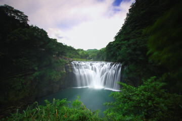 Fototapeta premium The breathtaking Shifen Waterfall plunges into an emerald pool, surrounded by lush green forests under a clear blue sky in Pingxi, New Taipei City, Taiwan, during summer.