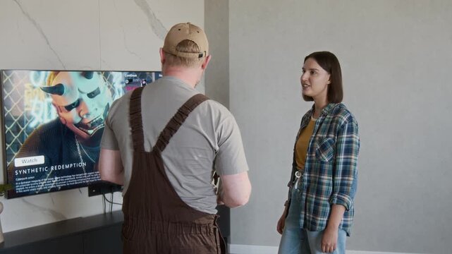 Medium shot of smiling young woman shaking hands with repairman standing by TV in home living room after successful home maintenance job