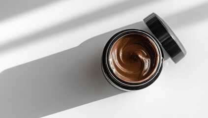 Dark brown cosmetic cream in a black jar.  Top-down view, showing the swirled texture of the product.  Shadows cast from sunlight or artificial light