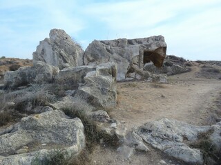 Ancient weathered rocks rise from a dusty trail under a pale blue sky, forming natural sculptures. The rugged terrain invites exploration, echoing stories of time, wind, and silent erosion.