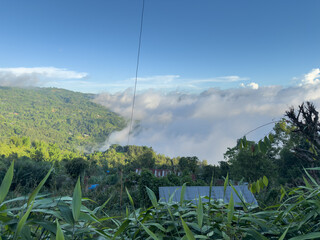 Scenic mountain landscape with winding rural road through green hills and lush vegetation under dramatic cloudy blue sky, peaceful countryside view with scattered houses and dense forest cover