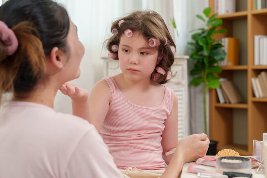 Asian daughter playfully holding powder puff while interacting with smiling mother during beauty session, strong emotional connection, nurturing trust and affection between family members at home.