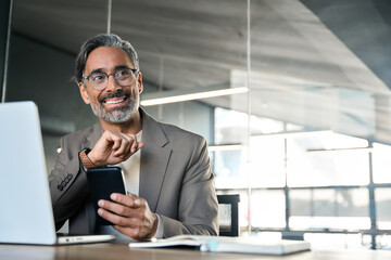 Mature businessman professional executive holding cellphone working at desk with laptop smartphone looking at copy space. Happy middle aged business man investor sitting in office using mobile phone.