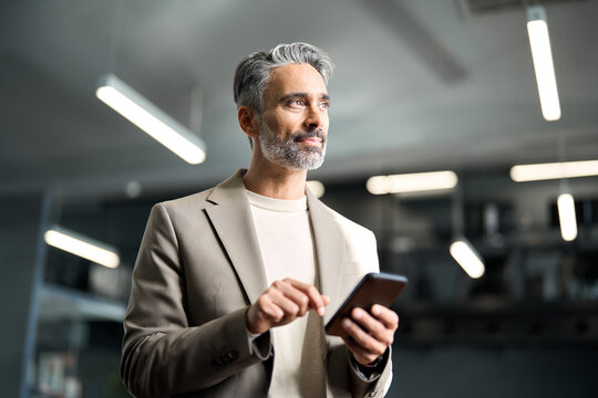 Mature older businessman executive leader wearing suit holding mobile phone at work standing with smartphone in hands. Middle aged business man wearing suit using cellphone looking away in office. - Powered by Adobe