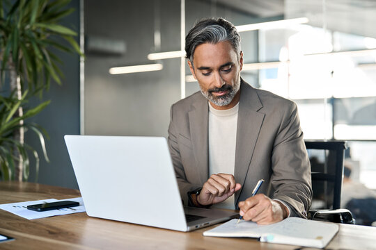 Middle aged professional executive manager lawyer, busy mature business man wearing suit writing notes watching online webinar analysing financial data using laptop computer working in office at desk.