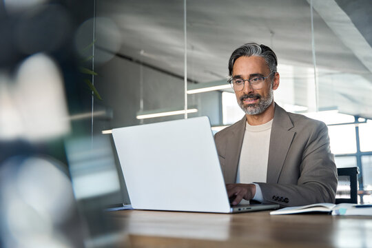 Professional busy middle aged Latin business man executive ceo wearing suit and glasses sitting at desk using laptop. Mature businessman manager working looking at computer technology in office.