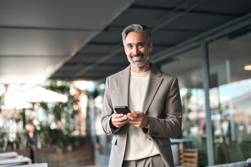 Happy middle aged businessman manager or entrepreneur holding smartphone looking at camera standing outside office. Mature older business man executive using mobile cell phone outdoors. Portrait.