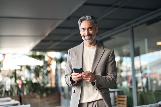 Happy middle aged businessman manager or entrepreneur holding smartphone looking at camera standing outside office. Mature older business man executive using mobile cell phone outdoors. Portrait. - Powered by Adobe