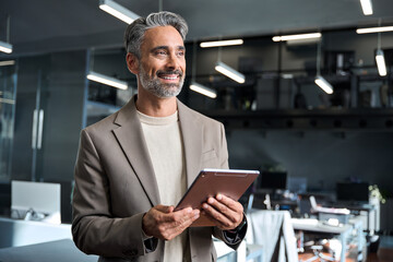 Smiling middle aged professional businessman manager wearing suit looking away at work in modern space. Happy confident mature business man executive standing in corporate office using digital tablet.