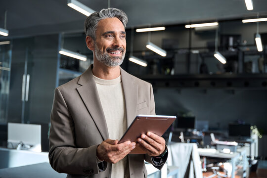 Smiling middle aged professional businessman manager wearing suit looking away at work in modern space. Happy confident mature business man executive standing in corporate office using digital tablet.
