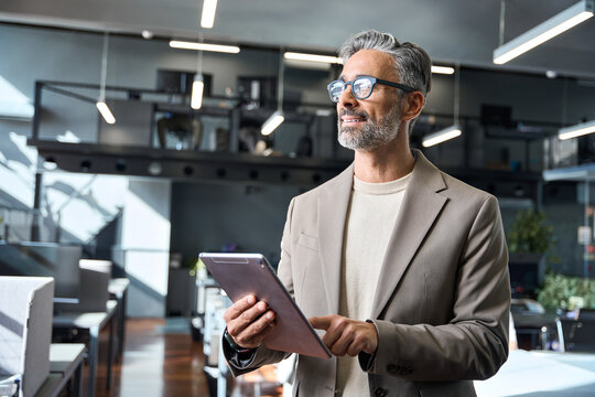 Busy confident mature 50 years old businessman ceo investor wearing glasses and suit thinking of future. Middle aged business man professional executive standing in office using tablet looking away.