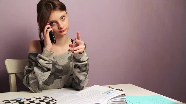 Fun seious emotion teen doing homework for school sitting at a desk holding pen in hand with book, copy books, pens, pencil case and talking on mobile phone. Back to school. Emotional education video
