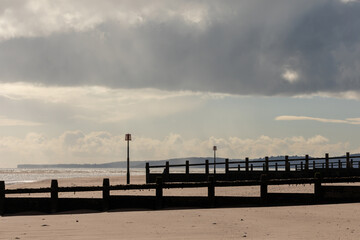 Obraz premium Moody sky over quiet sandy beach with wooden groynes and distant hills, capturing the calm and atmospheric beauty of the British coastline.