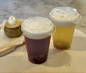 Two plastic cups with fruit bubble tea and a cake in the background, on a wooden table