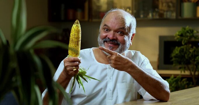 Senior Indian man eating roasted maize or bhutta at home in modern kitchen, Asian foodie old man expressing immense joy and happiness while enjoying healthy traditional Indian snack at dining table