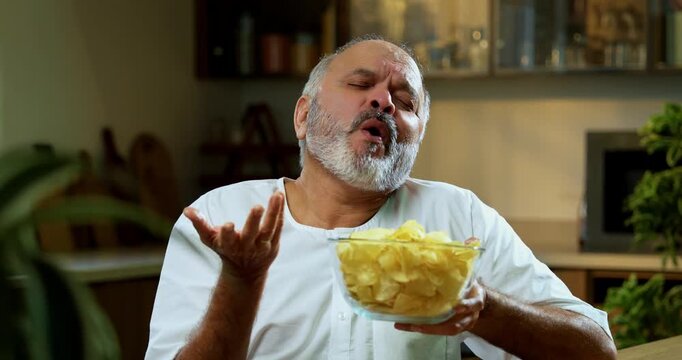 Senior Indian man eating potato chips secretly at home in modern kitchen, Asian foodie old man expressing immense happiness while enjoying unhealthy wafers snack alone at dining table