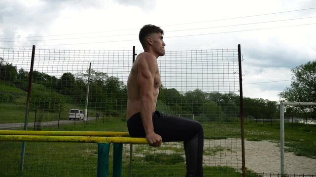 Strong guy training abdominal muscles on parallel bars at sports ground. Young shirtless sportsman exercising at nature. Sporty athlete working out at nature. Concept of active sportive lifestyle