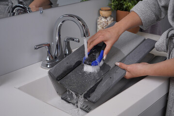 A person scrubs a gray slat bath mat with a blue and white brush under running water in a bathroom sink. The mat's foldable design makes it easy to clean