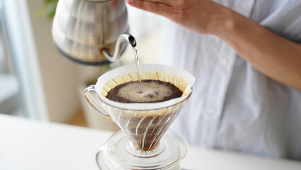 Close-up of a person's hand pouring hot water from a gooseneck kettle, brewing fresh pour-over coffee in a glass dripper with bright natural light.
