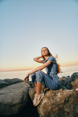 young woman sitting on rocks by the calm sea at sunset, wearing casual denim outfit and sneakers, relaxed outdoor lifestyle with natural lighting