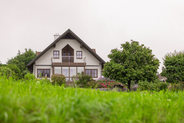 A charming two-story house with a steep roof and balcony sits amidst lush greenery. The foreground features tall grass, while trees and colorful flowers surround the home under a cloudy sky.