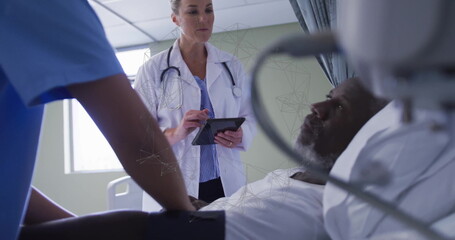 Consulting doctor tapping tablet in white coat at hospital bed, with nurse in scrubs adjusting arm