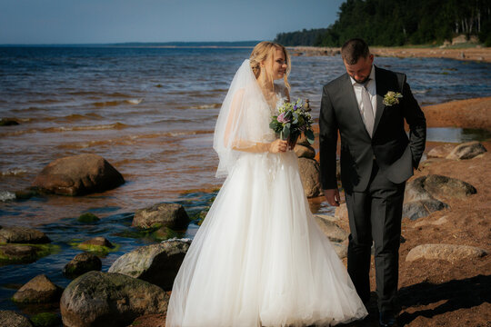 Groom kneels and gazes at the smiling bride standing on a rocky beach, surrounded by waves and a scenic shoreline. - Powered by Adobe