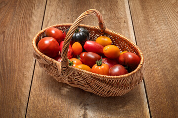 A wicker basket filled with colorful fresh tomatoes on a wooden background. A variety of types of tomatoes, including red, yellow, orange