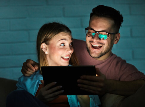 Portrait of young happy couple using tablet computer at night, using modern technology having fun with glowing screen in dark office or at home, watching tv together at home. Shot of a couple resting 