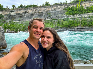 A young couple smiles while taking a selfie with the Niagara rapids and the Escarpment in the background.