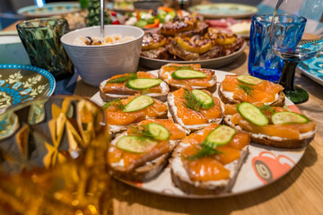 A close-up of a festive Christmas dinner spread on a wooden table. Homemade dishes, including open-faced sandwiches, a fresh salad, baked meat with cheese, and a platter of cold cuts and cheeses.