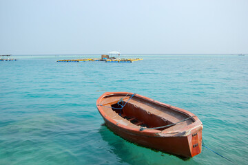 Rustic wooden rowboat moored on tranquil turquoise ocean with distant floating structures under hazy sky inviting exploration adventure summer travel destination relaxation