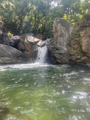Small waterfall cascading into a clear green pool, surrounded by rocks and tropical forest — a...