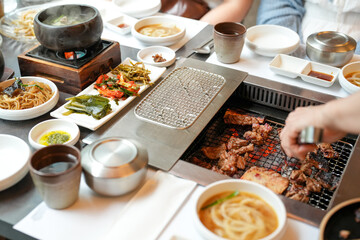 In a kalbi barbecue restaurant, a server grills a cut of meat over glowing coals, flames licking edges as steam rises from sizzling fat, filling the air with rich aroma. Seoul, Jung District, spring.