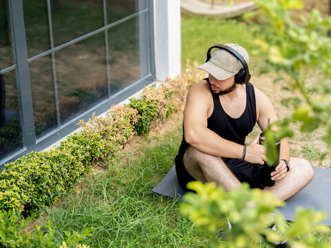 Young man working out outdoors, doing bodyweight exercises, checking fitness watch, hydrating, and relaxing. Wearing headphones in a garden setting. Symbol of health and active lifestyle. - Powered by Adobe