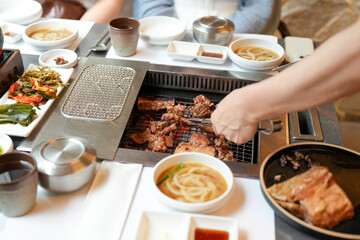 In a kalbi barbecue restaurant, a server grills a cut of meat over glowing coals, flames licking edges as steam rises from sizzling fat, filling the air with rich aroma. Seoul, Jung District, spring.