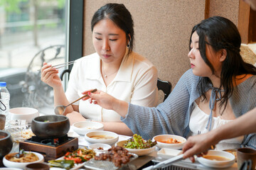 In a kalbi barbecue restaurant at a sunlit window table, two Korean women in their twenties in casual attire enjoy grilled meat and side dishes under soft spring light. Seoul, Jung District, spring.