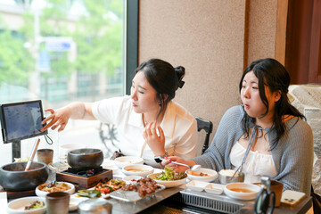 In a kalbi barbecue restaurant at a sunlit window table, two Korean women in their twenties in casual attire enjoy grilled meat and side dishes under soft spring light. Seoul, Jung District, spring.