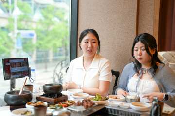 In a kalbi barbecue restaurant at a sunlit window table, two Korean women in their twenties in casual attire enjoy grilled meat and side dishes under soft spring light. Seoul, Jung District, spring.