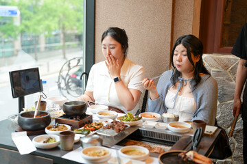 In a kalbi barbecue restaurant at a sunlit window table, two Korean women in their twenties in casual attire enjoy grilled meat and side dishes under soft spring light. Seoul, Jung District, spring.