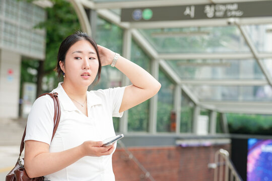 A Korean woman couple in their twenties in casual clothes stand by a stairway entrance to the subway, talking and using a smartphone, sharing plans under spring light. Seoul, Jung District, spring.