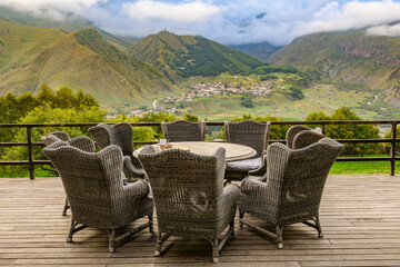 Fototapeta premium Elevated deck in Stepantsminda, with a view of the Caucasus and Gergeti Trinity Church. Wicker chairs surround a round table with refreshments.