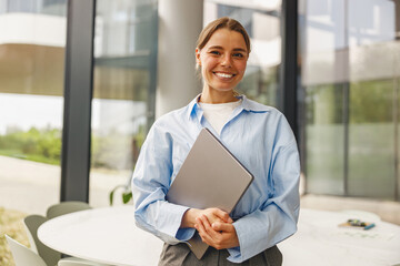 A cheerful and professional woman holding a laptop stands confidently in a bright, contemporary office setting © Yaroslav Astakhov