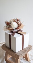 Dried floral bouquet atop a large, white, gift box tied with brown ribbon, sitting on a rustic wooden stool against a plain backdrop