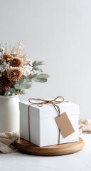 A minimalist still life featuring a white gift box tied with twine and a blank tag, resting on a wooden round, beside a vase of dried flowers on a white surface