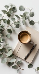 Flatlay of espresso in a dark mug atop a beige notebook, alongside eucalyptus sprigs and a pen on a white background