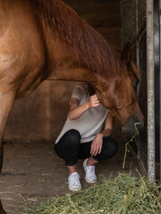Young person crouching beside chestnut horse in stable showing intimate human-animal connection and...